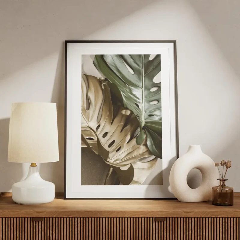 affiche de feuilles de monstera une verte et une doree sur un fond beige avec un meuble.webp